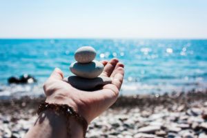 A hand holds a tower of three smooth rocks on one another in front of the ocean. This could represent the peace an online therapist in Michigan can help you achieve. Contact a young adult therapist in West Bloomfield, MI to learn more today! 48323 | 94118 | 48322
